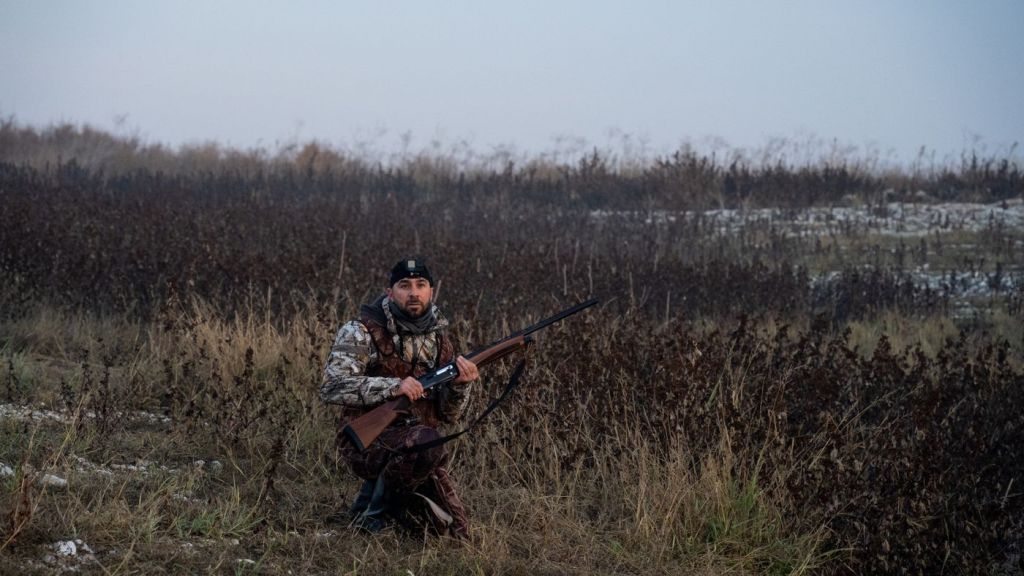 A hunter kneeling in tall grass holding a shotgun