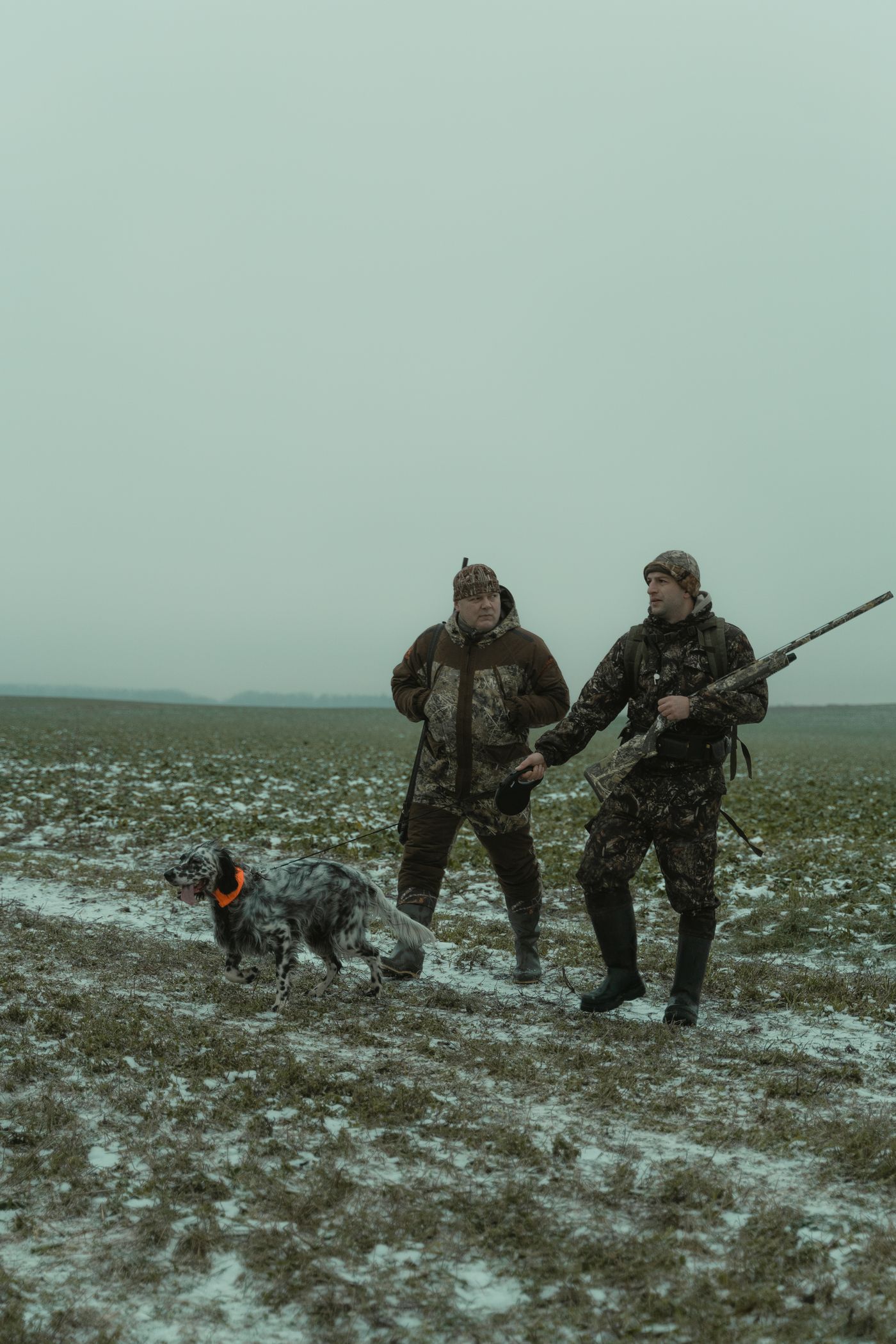 Two hunters with shotguns walking a field with a dog