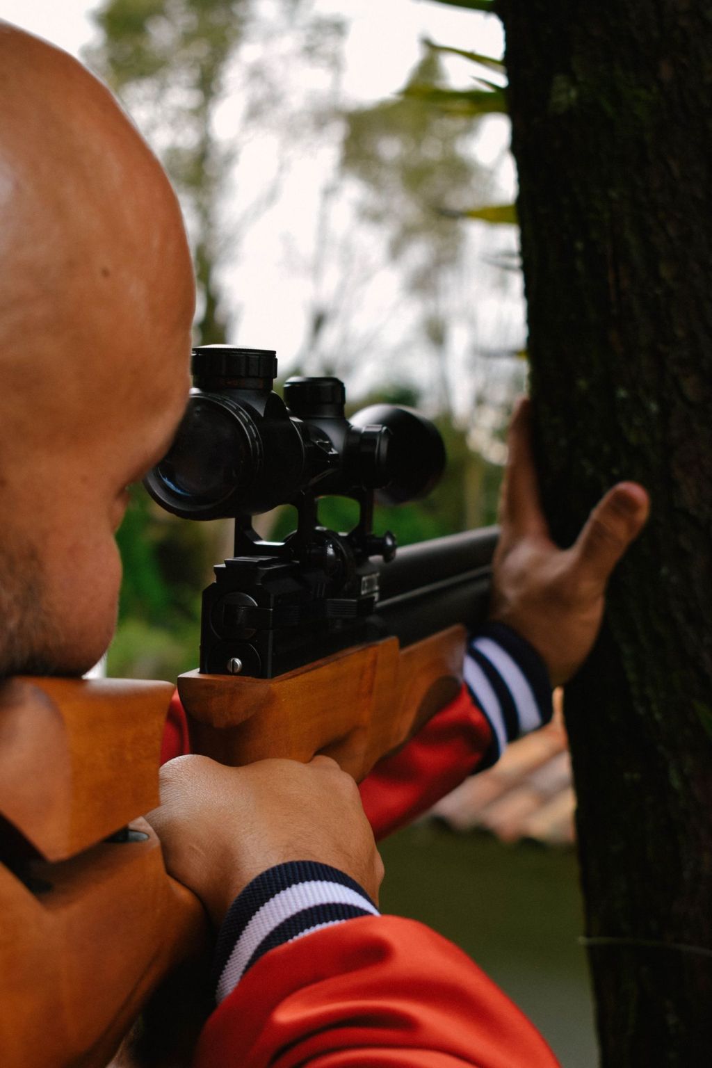 A hunter aiming a scoped rifle from beside a tree
