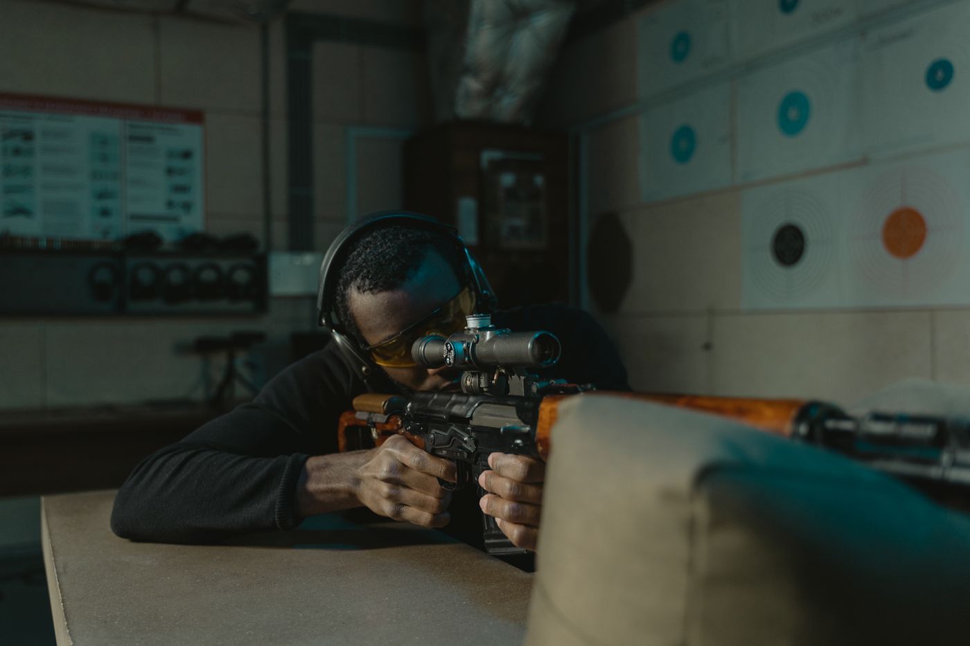A shooter wearing eye and ear protection aiming a rifle at an indoor range