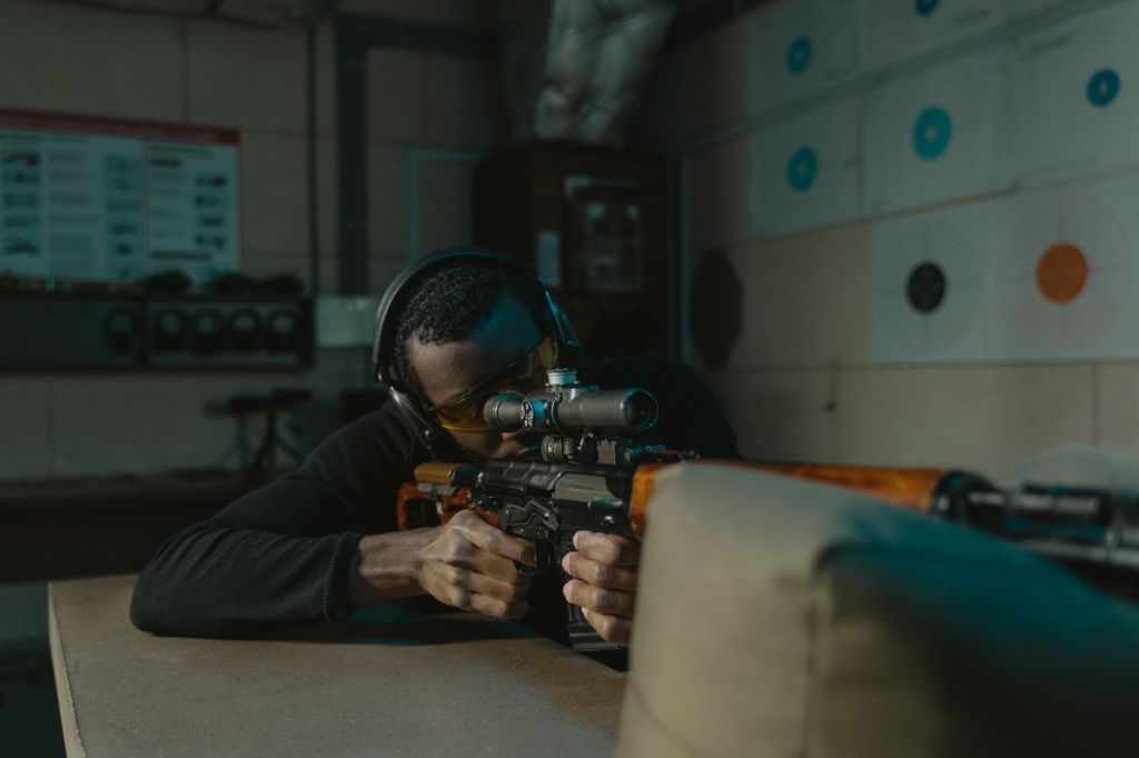 A shooter wearing eye and ear protection aiming a rifle at an indoor range