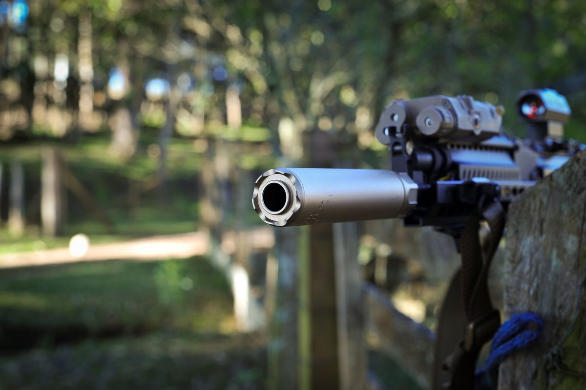Close-up of a rifle fitted with a suppressor, photographed outdoors near a wooden fence